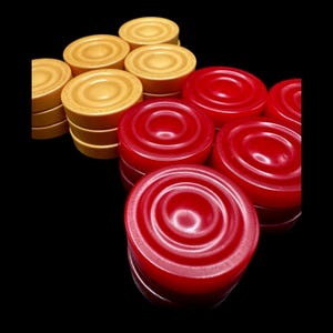 May include: A collection of red and tan checkers, with a glossy finish, arranged against a black background. The checkers are round with concentric circles on the top surface. The tan checkers are stacked in groups, while the red checkers are scattered.