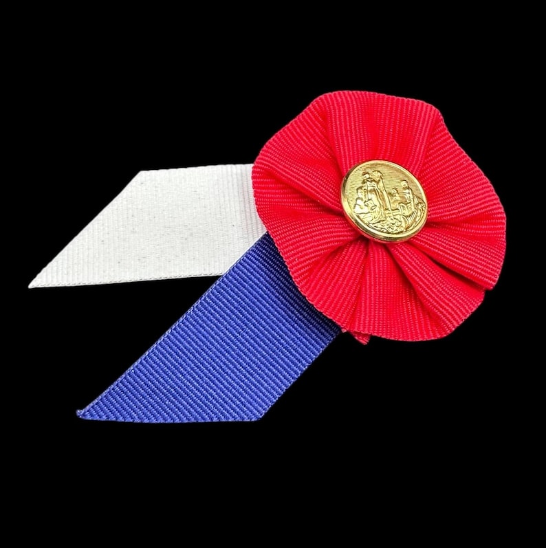 May include: A patriotic rosette with a red, white, and blue color scheme. The red ribbon is gathered into a circular shape, with a gold-colored button in the center. A white and blue ribbon extends from the rosette.