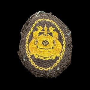 May include: A brown fabric patch with a gold embroidered diving helmet and trident design. The helmet is surrounded by a chain link design.