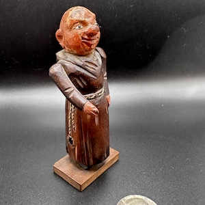 May include: A hand-carved wooden figurine of a monk with a brown robe and a rope belt. The monk has a smiling face with a large nose and a white beard.