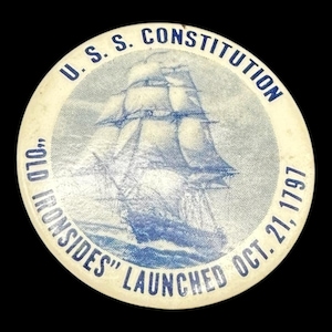 May include: A round, off-white button with a blue illustration of a tall ship sailing on the ocean. The text "U.S.S. CONSTITUTION" arches across the top, with "OLD IRONSIDES" and "LAUNCHED OCT. 21, 1797" in a circular pattern.