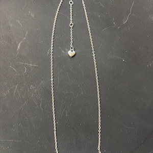 May include: A silver-toned necklace featuring a thin chain and a rectangular bar pendant with an embossed swirl pattern. The necklace includes an adjustable chain section ending in a small heart-shaped charm with the letters OB engraved on it.