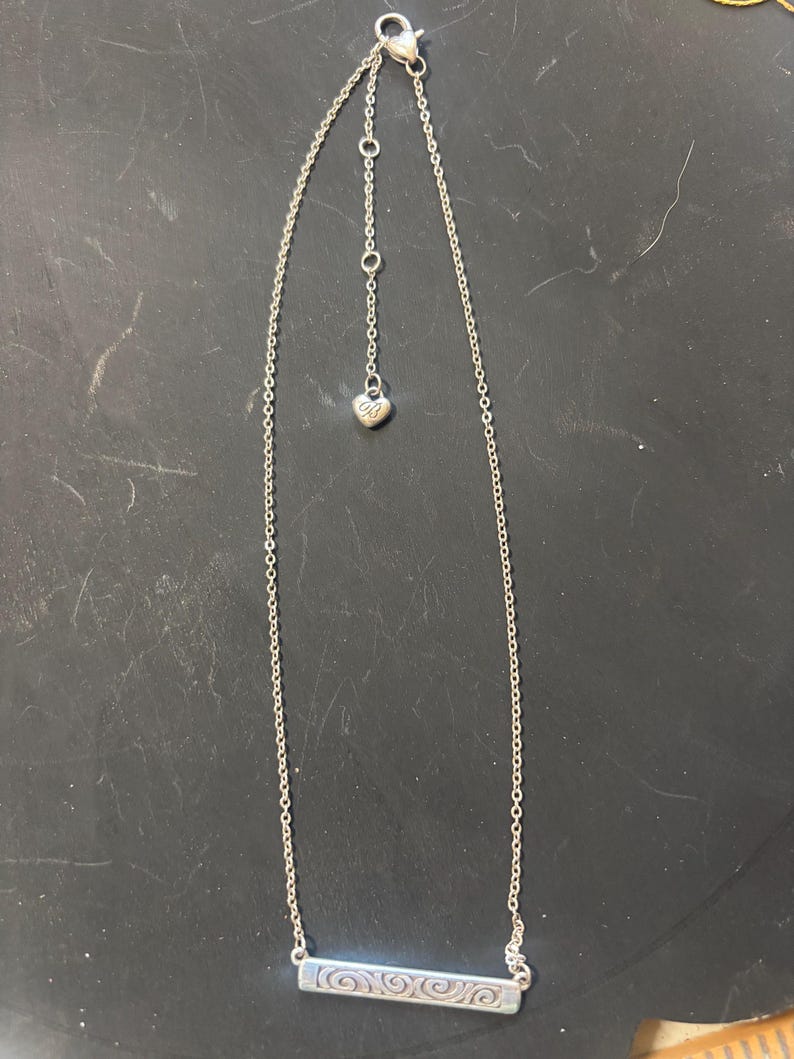 May include: A silver-toned necklace featuring a thin chain and a rectangular bar pendant with an embossed swirl pattern. The necklace includes an adjustable chain section ending in a small heart-shaped charm with the letters OB engraved on it.