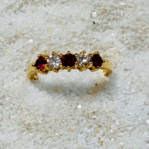 May include: A gold ring featuring five round-cut gemstones arranged in a row. The stones alternate between three deep red garnets and two clear white diamonds, all set in a polished gold band with prong settings holding each individual stone.