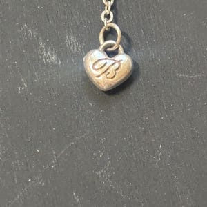 May include: A silver-toned metal chain necklace featuring a small, heart-shaped charm. The charm is embossed with the capital letter B in a cursive script font. The pendant is attached to the chain by a small circular jump ring.