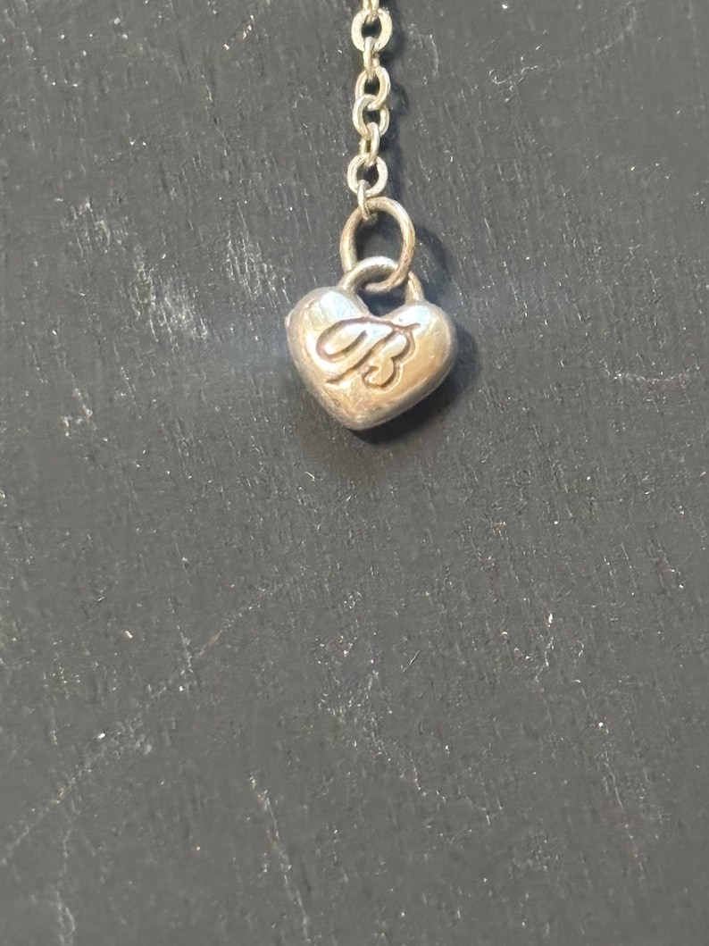 May include: A silver-toned metal chain necklace featuring a small, heart-shaped charm. The charm is embossed with the capital letter B in a cursive script font. The pendant is attached to the chain by a small circular jump ring.