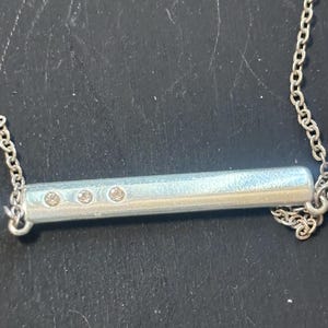 May include: A silver-toned horizontal bar pendant necklace featuring a smooth, cylindrical metal bar with three small, clear, circular inset stones arranged in a row on the left side, suspended from a delicate silver-toned link chain.