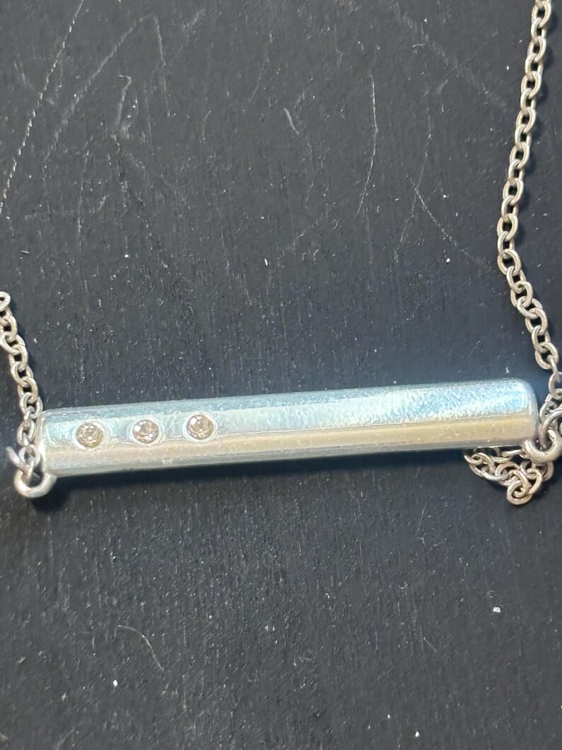 May include: A silver-toned horizontal bar pendant necklace featuring a smooth, cylindrical metal bar with three small, clear, circular inset stones arranged in a row on the left side, suspended from a delicate silver-toned link chain.