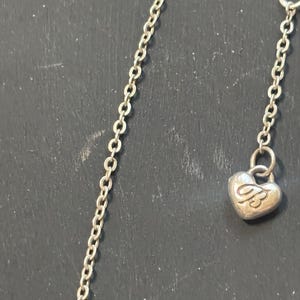 May include: A silver-toned metal chain necklace featuring a small, heart-shaped charm dangling from the end. The charm is embossed with the letters JB in a stylized script font. The chain consists of small, uniform oval links connected in a series.
