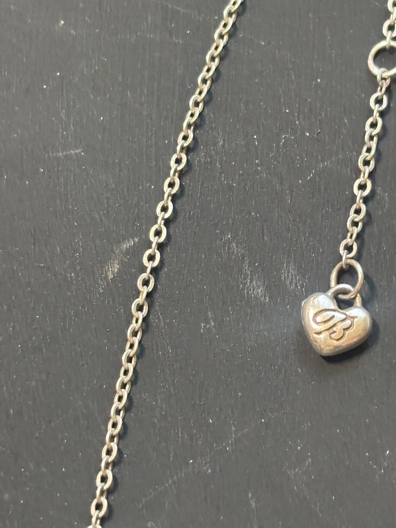 May include: A silver-toned metal chain necklace featuring a small, heart-shaped charm dangling from the end. The charm is embossed with the letters JB in a stylized script font. The chain consists of small, uniform oval links connected in a series.