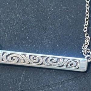 May include: A silver-toned metal bar pendant necklace featuring a series of embossed, repeating spiral patterns along the length of the rectangular bar. The pendant is attached to a delicate, thin silver-toned metal chain link necklace.