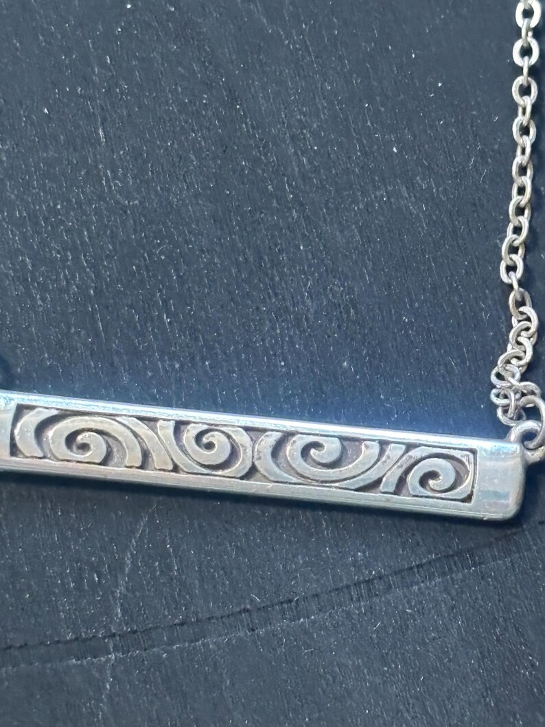 May include: A silver-toned metal bar pendant necklace featuring a series of embossed, repeating spiral patterns along the length of the rectangular bar. The pendant is attached to a delicate, thin silver-toned metal chain link necklace.