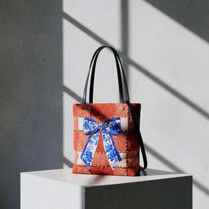 May include: A red and white patchwork tote bag with a large blue floral bow. The bag has black handles and a small gold button detail. The bag is sitting on a white cube.