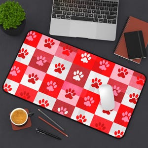May include: A rectangular mouse mat featuring a red, white, and pink chequered design. The pattern is adorned with paw prints in shades of red and pink. A white computer mouse rests on the mat.