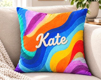 May include: A square throw pillow featuring a vibrant, multi-colored wavy pattern in shades of blue, green, yellow, orange, red, and purple. The name Kate is embroidered in white, textured, raised script lettering across the center of the pillow.