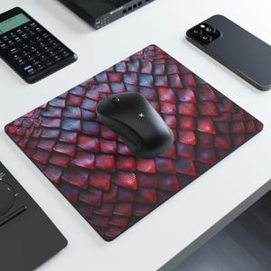 May include: A black wireless computer mouse sits on a rectangular mousepad. The mousepad has a red and purple scale pattern. Other items on the white surface include a calculator, a phone, and a laptop.
