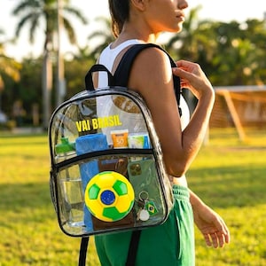 Puede incluir: Una mochila de plástico transparente con correas y bordes negros, que presenta el texto amarillo VAI BRASIL impreso en el frente. La bolsa contiene una toalla azul, una botella de agua verde, un pequeño balón de fútbol y un teléfono inteligente.