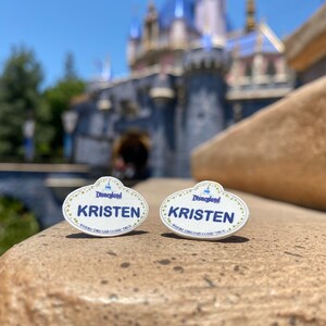 K&ouml;nnte beinhalten: Zwei wei&szlig;e Disneyland-Namensschilder mit goldenen Verzierungen und blauen Buchstaben. Beide Schilder zeigen "Disneyland" und "KRISTEN" mit dem Slogan "Where Dreams Come True".