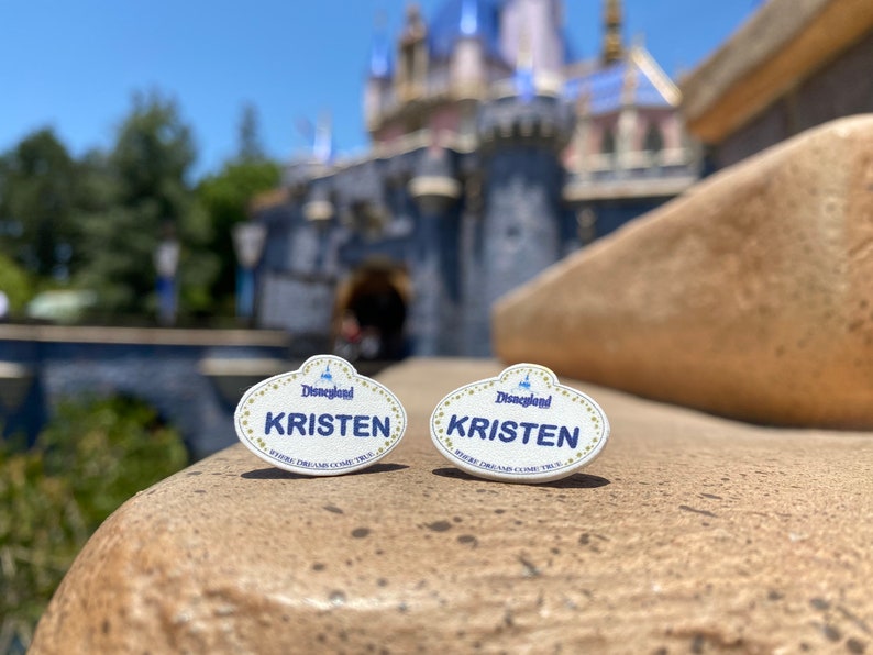 K&ouml;nnte beinhalten: Zwei wei&szlig;e Disneyland-Namensschilder mit goldenen Verzierungen und blauen Buchstaben. Beide Schilder zeigen "Disneyland" und "KRISTEN" mit dem Slogan "Where Dreams Come True".