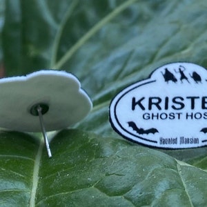 K&ouml;nnte beinhalten: Ein Paar wei&szlig;e und schwarze Ohrringe zum Thema Haunted Mansion. Die Ohrringe zeigen ein Namensschild mit dem Text "Kristen Ghost Host" und den Worten "Haunted Mansion".