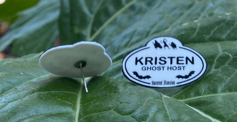K&ouml;nnte beinhalten: Ein Paar wei&szlig;e und schwarze Ohrringe zum Thema Haunted Mansion. Die Ohrringe zeigen ein Namensschild mit dem Text "Kristen Ghost Host" und den Worten "Haunted Mansion".