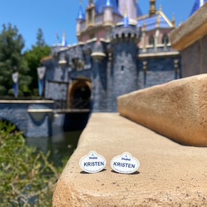 K&ouml;nnte beinhalten: Zwei wei&szlig;e Disneyland-Namensschilder mit blauer Beschriftung. Die Pins zeigen "Disneyland" und "KRISTEN". Die Pins liegen auf einer braunen Steinmauer vor einem verschwommenen Bild von Dornr&ouml;schens Schloss.