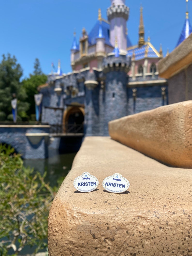 K&ouml;nnte beinhalten: Zwei wei&szlig;e Disneyland-Namensschilder mit blauer Beschriftung. Die Pins zeigen "Disneyland" und "KRISTEN". Die Pins liegen auf einer braunen Steinmauer vor einem verschwommenen Bild von Dornr&ouml;schens Schloss.