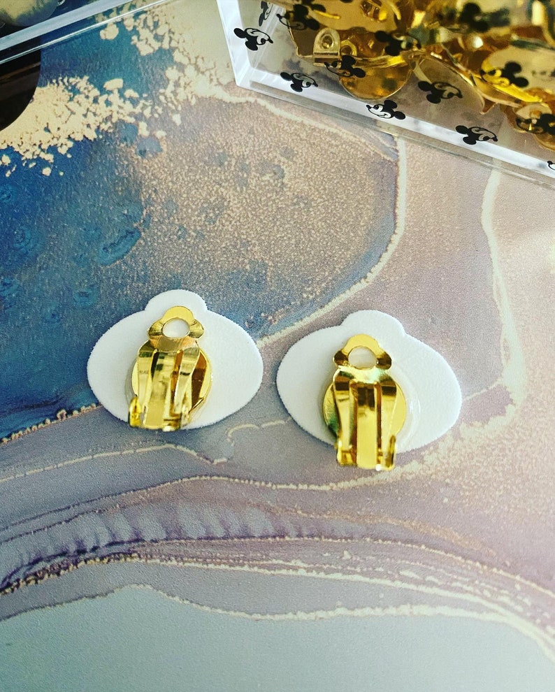 K&ouml;nnte beinhalten: Ein Paar goldfarbene Ohrclips mit wei&szlig;er R&uuml;ckseite. Die Clips sind oval geformt und haben eine glatte Oberfl&auml;che.