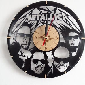 Puede incluir: Un reloj de vinilo negro con la banda Metallica. La esfera del reloj es un círculo de madera con agujas negras y números romanos. El reloj está decorado con el logotipo de la banda y cuatro miembros de la banda.