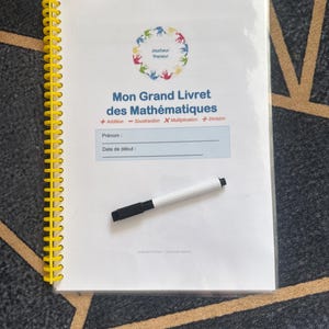 Könnte beinhalten: Ein weißes Mathematik-Arbeitsheft mit gelber Spiralbindung aus Kunststoff. Auf dem Einband steht "Mon Grand Livret des Mathématiques" sowie ein kreisförmiges Logo mit bunten Handabdrücken. Ein schwarzer Whiteboard-Marker liegt auf dem Heft.