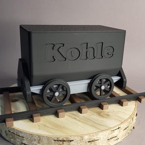 Könnte beinhalten: Ein schwarzer Spielzeug-Kohlewagen mit dem Wort "Kohle" auf der Seite, der auf einer Holzbasis mit einer Schiene steht. Der Wagen hat vier Räder und ist so gestaltet, dass er einem Miniatur-Kohlewagen ähnelt.