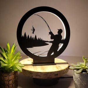 Könnte beinhalten: Schwarze Metalllampe mit einer Fischerszene in einem kreisförmigen Rahmen. Die Silhouette zeigt einen Fischer, der einen Fisch einholt, mit einem Wald und einem Fluss im Hintergrund. Die Lampe steht auf einem Holzsockel.