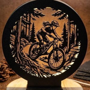 Könnte beinhalten: Schwarze, kreisförmige, lasergeschnittene Holzlampe mit einem Mountainbiker auf einem Trail. Das Design umfasst Bäume und eine Waldszene, mit warmem Licht im Inneren. Die Lampe steht auf einem Holzsockel.