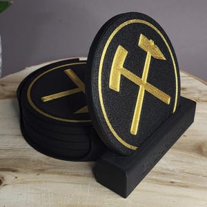 Könnte beinhalten: Ein Satz schwarzer Untersetzer mit einem goldenen Emblem gekreuzter Hämmer, präsentiert in einem schwarzen Halter. Die Untersetzer sind rund und haben eine strukturierte Oberfläche. Das Design ist ein goldenes Emblem auf schwarzem Hintergrund.