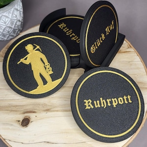 Könnte beinhalten: Set aus vier schwarzen Untersetzern mit goldenen Akzenten. Ein Untersetzer zeigt eine Bergmann-Silhouette, während andere die Wörter "Ruhrpott" und "Glück Auf" in goldener Schrift zeigen. Die Untersetzer werden auf einer Holzoberfläche präsentiert.