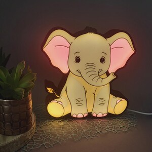 Könnte beinhalten: Niedliches Nachtlicht in Form eines Elefanten im Cartoon-Stil. Der Elefant ist hellgelb mit rosa Ohren und einem lächelnden Gesicht. Das Licht hat eine schwarze Umrandung und steht auf einem Tisch mit einer Pflanze.