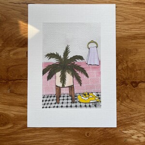 May include: A framed art print depicting a bathroom scene. A potted palm sits on a wooden stand. A yellow pool float with the text "MARZ" is on the black and white tiled floor. A pink brick wall and a towel on a gold ring are in the background.
