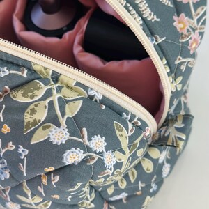 May include: A quilted cosmetic bag with a floral pattern in shades of teal, green, and pink. The bag is open, revealing a pink interior and compartments holding makeup items. The zipper is a cream color.