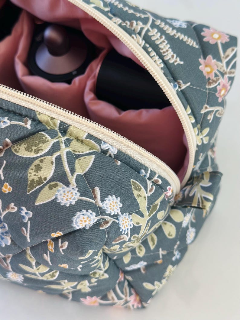 May include: A quilted cosmetic bag with a floral pattern in shades of teal, green, and pink. The bag is open, revealing a pink interior and compartments holding makeup items. The zipper is a cream color.