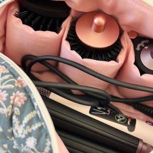 May include: A floral-patterned cosmetic bag with a pink interior, holding hair styling tools. The bag contains round brushes, a curling iron, and a flat iron. The bag is blue with a white and pink floral design.