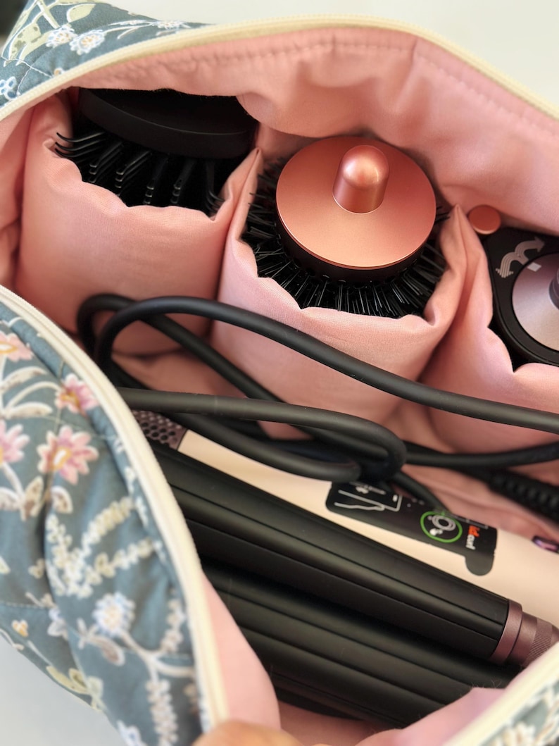 May include: A floral-patterned cosmetic bag with a pink interior, holding hair styling tools. The bag contains round brushes, a curling iron, and a flat iron. The bag is blue with a white and pink floral design.