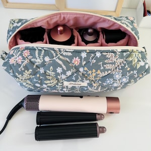May include: A floral-patterned quilted cosmetic bag with a pink interior, open to reveal compartments holding beauty tools. A hair dryer and two curling iron attachments are in front of the bag. The bag has the word "cloudesign" on it.