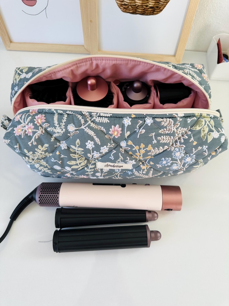 May include: A floral-patterned quilted cosmetic bag with a pink interior, open to reveal compartments holding beauty tools. A hair dryer and two curling iron attachments are in front of the bag. The bag has the word "cloudesign" on it.