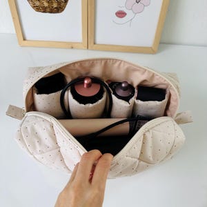 Può includere: Un beauty case beige trapuntato con pois neri, aperto per rivelare scomparti e strumenti di bellezza. La borsa ha un interno rosa chiaro e una chiusura con cerniera. La borsa è su una superficie bianca.