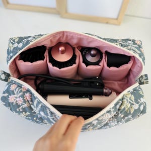 May include: A floral-patterned cosmetic bag in shades of gray, pink, and white, open to reveal hair styling tools. The bag contains a hair straightener, brushes, and other beauty accessories, all neatly organized within.