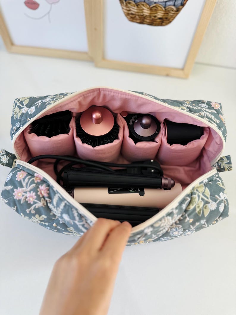 May include: A floral-patterned cosmetic bag in shades of gray, pink, and white, open to reveal hair styling tools. The bag contains a hair straightener, brushes, and other beauty accessories, all neatly organized within.
