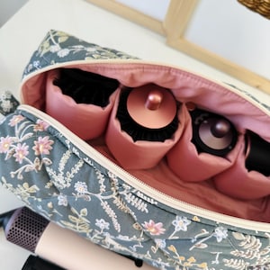 May include: A floral-patterned travel case with a pink interior, holding hair styling tools. The case is teal with a quilted design and features a zipper closure. Hair styling tools include a hair dryer and various brush attachments.