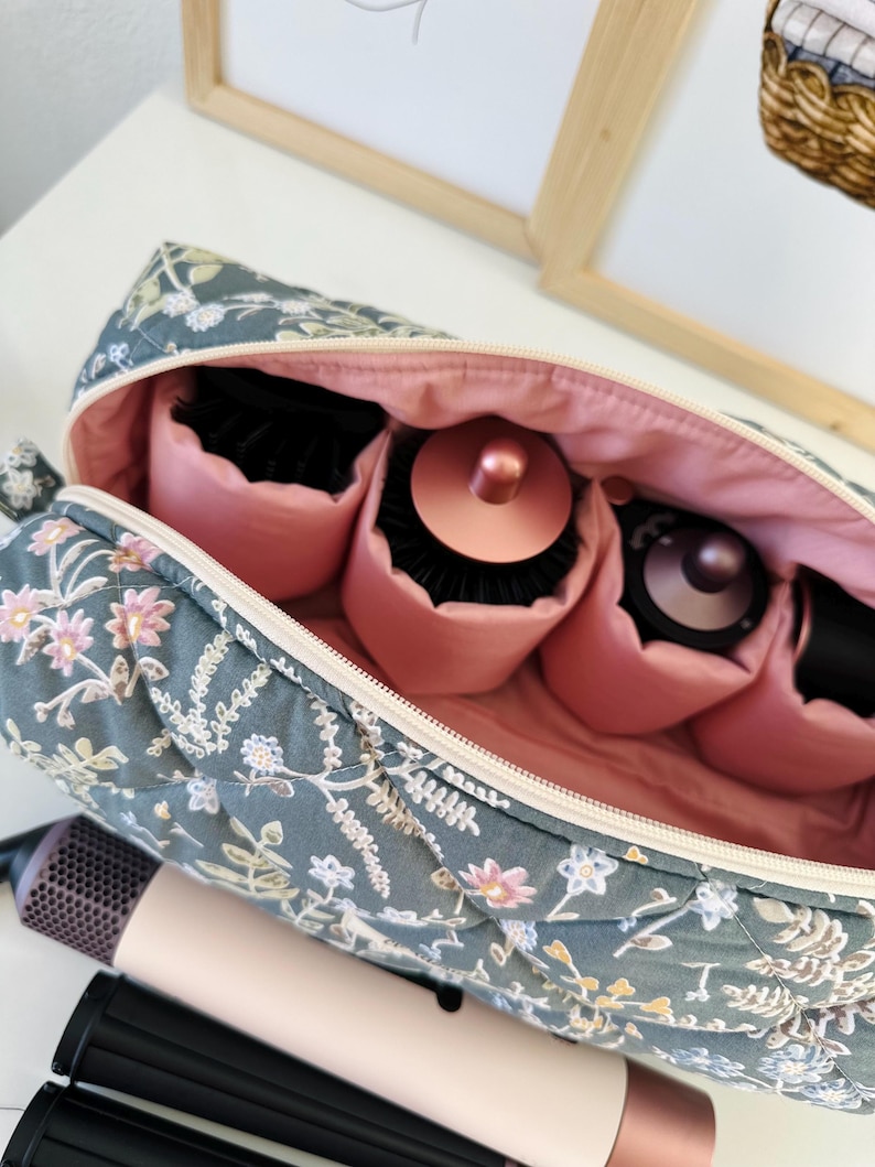 May include: A floral-patterned travel case with a pink interior, holding hair styling tools. The case is teal with a quilted design and features a zipper closure. Hair styling tools include a hair dryer and various brush attachments.