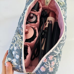May include: A floral-patterned quilted cosmetic bag, open to reveal various hair styling tools. The bag is gray with a pink interior and a zipper closure. The bag contains a hair dryer, curling iron, and brushes.