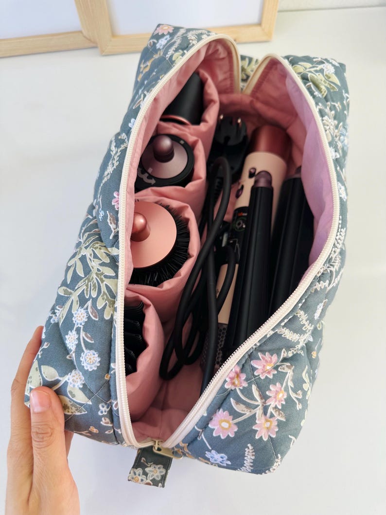 May include: A floral-patterned quilted cosmetic bag, open to reveal various hair styling tools. The bag is gray with a pink interior and a zipper closure. The bag contains a hair dryer, curling iron, and brushes.